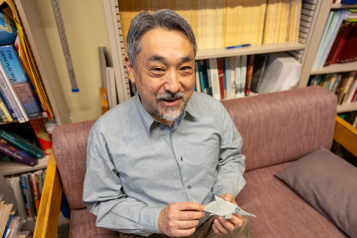 Motani sits on a couch in his office while holding a origami ichthyosaur.
