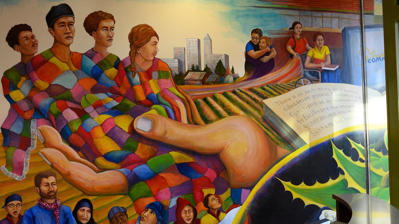 Multicolored wall mural of 4 diverse humans with a multicolored blanket draped over them, held by a large hand. Also green agricultural fields and workers nearby.